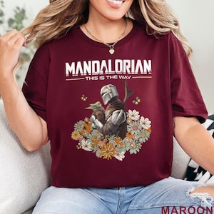 May include: Maroon t-shirt featuring the text "MANDALORIAN" above an illustration of the Mandalorian and Grogu surrounded by flowers. The text "THIS IS THE WAY" is below the main text. The word "MAROON" is at the bottom.