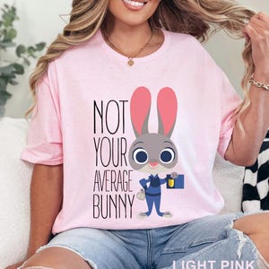 May include: Light pink t-shirt with the text "NOT YOUR AVERAGE BUNNY" and a cartoon bunny character wearing a blue uniform and holding a briefcase. The shirt is a crew neck style.