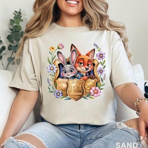May include: Sand-colored t-shirt featuring a colorful graphic of Judy Hopps and Nick Wilde from Zootopia, framed by flowers and police badges. The word "SAND" is printed at the bottom of the shirt.
