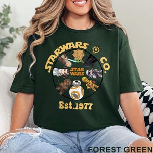 May include: Forest green t-shirt featuring a circular graphic with Star Wars characters and the text "STAR WARS CO. Est. 1977". The design includes characters like Yoda, Darth Vader, and BB-8. The shirt is a crew neck.