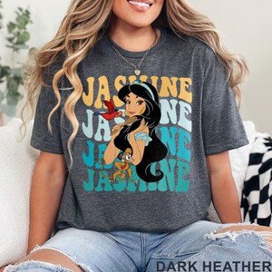May include: Dark heather gray t-shirt featuring a graphic of Princess Jasmine with the text "Jasmine" in a retro font. The design includes a parrot and a monkey. The shirt is made of a soft material.