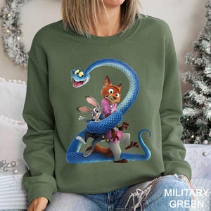 Puede incluir: Sudadera verde militar con un gr&aacute;fico de dibujos animados de una serpiente azul enrollada alrededor de un zorro y un conejo. El zorro lleva una chaqueta rosa y el conejo un traje. La sudadera es de color s&oacute;lido.