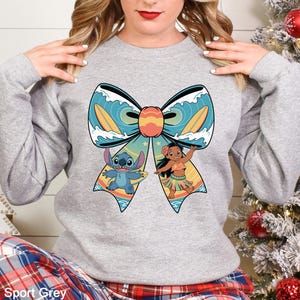 Puede incluir: Sudadera gris deportivo con un gran dise&ntilde;o de lazo con los personajes de dibujos animados Stitch y Lilo. El lazo tiene un fondo turquesa con tablas de surf y olas. Stitch sostiene una tabla de surf y Lilo baila.