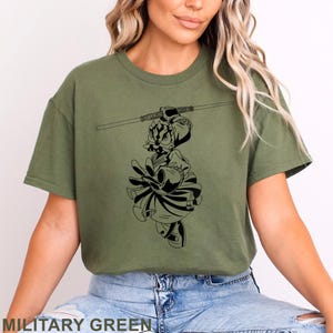 May include: Military green t-shirt featuring a black graphic of a cartoon character holding a lightsaber. The character is dressed in a warrior outfit. The text "MILITARY GREEN" is at the bottom of the image.