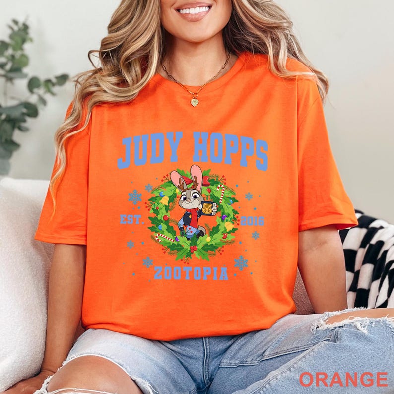 以下が含まれることがあります： オレンジ色のTシャツ。青色の文字で「JUDY HOPPS」と書かれており、ズートピアのジュディ・ホップスの漫画の画像がリースの中に描かれています。シャツには「EST. 2016」と「ZOOTOPIA」の文字も青色で書かれています。