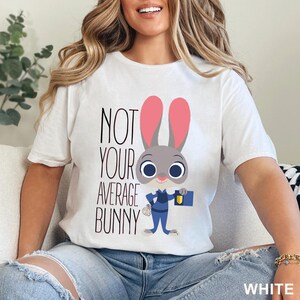 Puede incluir: Camiseta blanca con el texto "NOT YOUR AVERAGE BUNNY" y un dibujo de un conejo. El conejo tiene pelo gris, orejas rosas y lleva un uniforme azul. La camiseta es de un material suave.