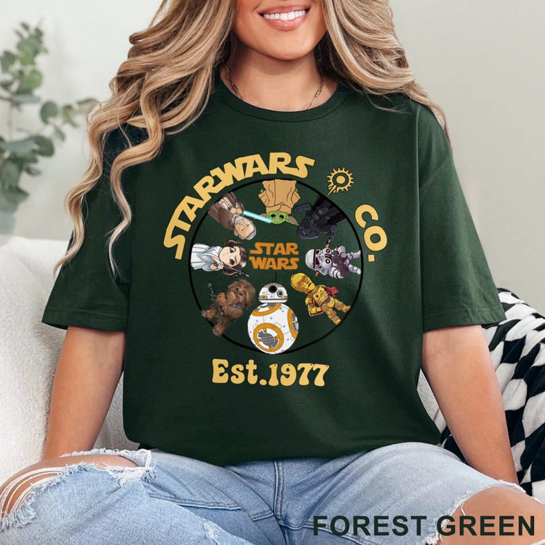 May include: Forest green t-shirt featuring a circular graphic with Star Wars characters and the text "STAR WARS CO. Est. 1977". The design includes characters like Yoda, Darth Vader, and BB-8. The shirt is a crew neck.
