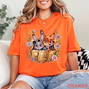 Puede incluir: Camiseta naranja brillante con un gr&aacute;fico de Judy Hopps y Nick Wilde de Zootopia, enmarcado por flores y placas de polic&iacute;a. La palabra "ORANGE" est&aacute; impresa en la parte inferior de la camiseta.