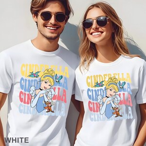 May include: White t-shirts featuring a colorful Cinderella graphic. The design includes the word "Cinderella" in stacked, retro-style lettering, along with an illustration of Cinderella and birds. The word "WHITE" is at the bottom.