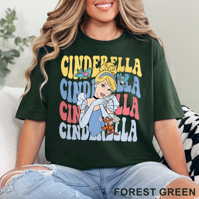 May include: Forest green t-shirt featuring a retro design with the word "Cinderella" stacked in yellow, blue, and pink. The graphic includes an illustration of Cinderella, birds, and her animal friends. The shirt is a comfortable, casual style.