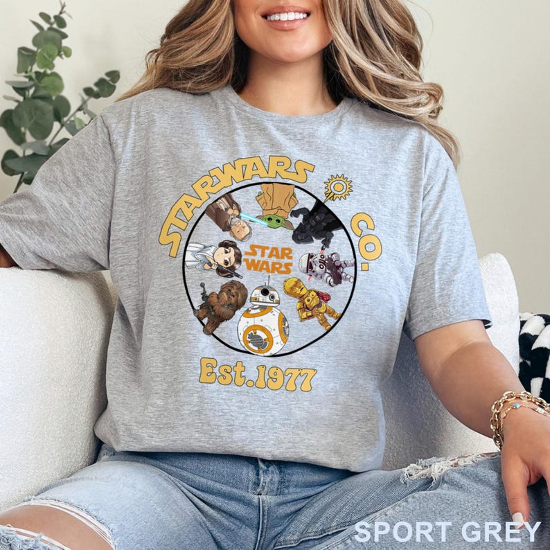 Peut inclure: T-shirt gris sport avec un motif circulaire repr&eacute;sentant des personnages de Star Wars et le texte "STAR WARS CO." et "Est. 1977" en jaune. Le t-shirt est de style col rond.