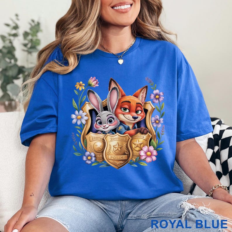 Puede incluir: Camiseta azul real con un gr&aacute;fico de Judy Hopps y Nick Wilde de Zootopia dentro de una placa de polic&iacute;a dorada, rodeada de flores. El texto "Zootopia Police" es visible en la placa. La camiseta es de color azul real liso.