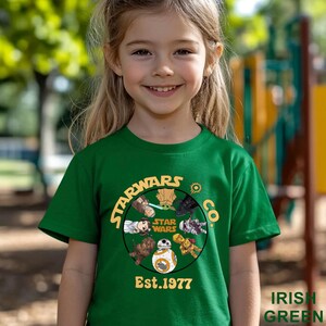 Peut inclure: Un t-shirt vert avec un motif Star Wars, comprenant des personnages et le texte "Est.1977". Le t-shirt est de couleur vert irlandais vif.