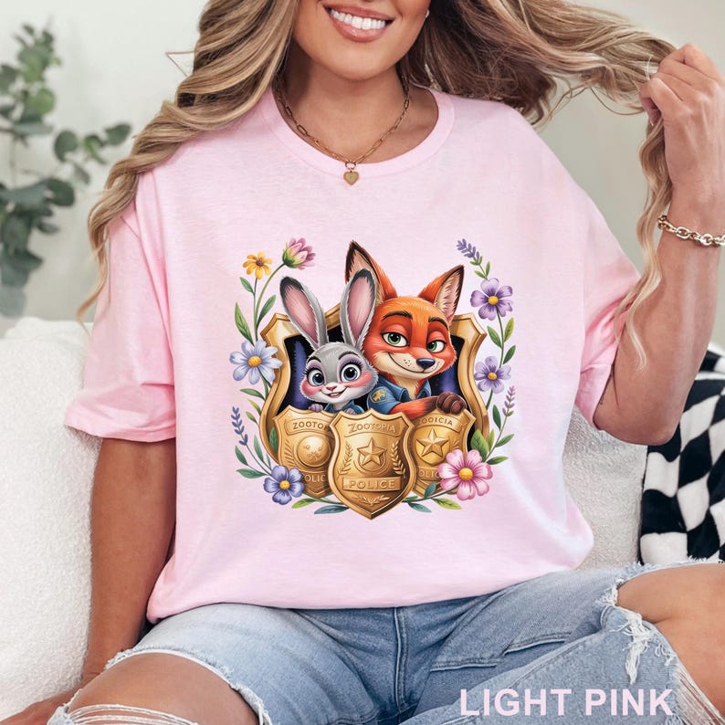 Puede incluir: Camiseta rosa claro con un gr&aacute;fico colorido de Judy Hopps y Nick Wilde de Zootopia dentro de una placa de polic&iacute;a, rodeada de flores. El texto "LIGHT PINK" est&aacute; en la parte inferior. Cuello redondo.