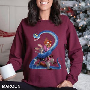 Puede incluir: Sudadera granate con una ilustraci&oacute;n de dibujos animados de una serpiente azul enrollada alrededor de un zorro y un conejo. El zorro lleva una chaqueta rosa y el conejo un traje. La palabra "MAROON" est&aacute; impresa en la parte inferior.