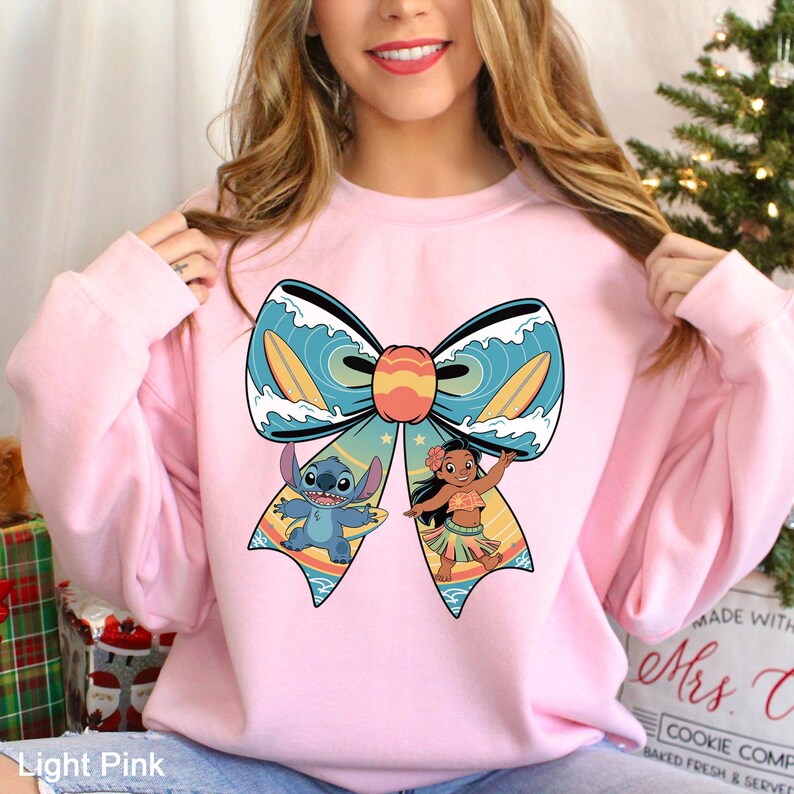 Puede incluir: Sudadera rosa claro con un gran dise&ntilde;o de lazo con los personajes de dibujos animados Stitch y Moana. El lazo tiene un patr&oacute;n de olas azules con tablas de surf amarillas. Stitch es azul y Moana lleva un top y una falda florales.