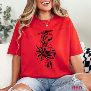Op de afbeelding: Rood T-shirt met een zwarte afbeelding van een personage met een staf. Het woord "RED" staat rechtsonder afgedrukt. Het shirt is effen rood en heeft korte mouwen.