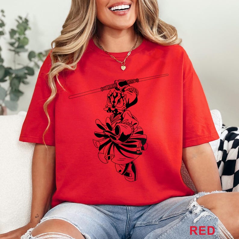 Op de afbeelding: Rood T-shirt met een zwarte afbeelding van een personage met een staf. Het woord "RED" staat rechtsonder afgedrukt. Het shirt is effen rood en heeft korte mouwen.