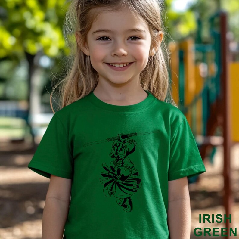 Op de afbeelding: Een groen t-shirt met een zwart grafisch ontwerp van een persoon op een scooter. De tekst "IRISH GREEN" is rechtsonder in het wit gedrukt. Het t-shirt wordt door een jong meisje gedragen.