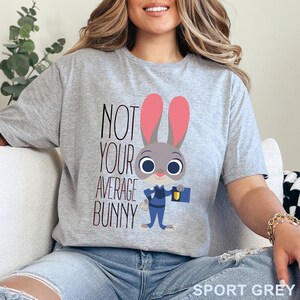 Puede incluir: Camiseta gris deportivo con un personaje de conejo de dibujos animados con uniforme azul, sosteniendo un malet&iacute;n y una bolsa de zanahorias. El texto "NOT YOUR AVERAGE BUNNY" est&aacute; impreso en la camiseta.