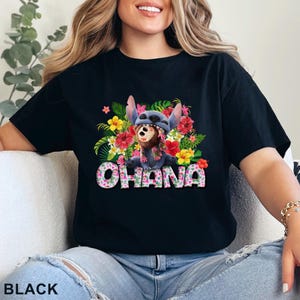 May include: Black t-shirt featuring a colorful graphic of a blue alien character with a floral lei and a brown bear. The word "OHANA" is printed in a floral pattern. The shirt is a crew neck.