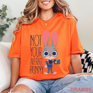 Puede incluir: Camiseta naranja brillante con el texto "NOT YOUR AVERAGE BUNNY" y un personaje de conejo de dibujos animados con uniforme de polic&iacute;a. El conejo tiene orejas grandes y lleva un malet&iacute;n. La palabra "ORANGE" est&aacute; en la parte inferior.
