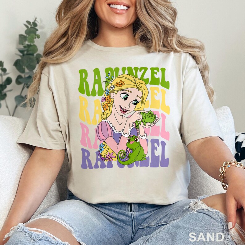 May include: Sand-colored t-shirt featuring a colorful graphic of Rapunzel holding a green chameleon. The word "Rapunzel" is stacked in green, yellow, pink, and purple. The shirt is a casual style.