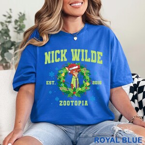 以下が含まれることがあります： ロイヤルブルーのTシャツ。緑色の「NICK WILDE」の文字の上に、クリスマスリースの中のニック・ワイルドの漫画の画像があります。シャツには「EST. 2016 ZOOTOPIA」の文字も含まれています。