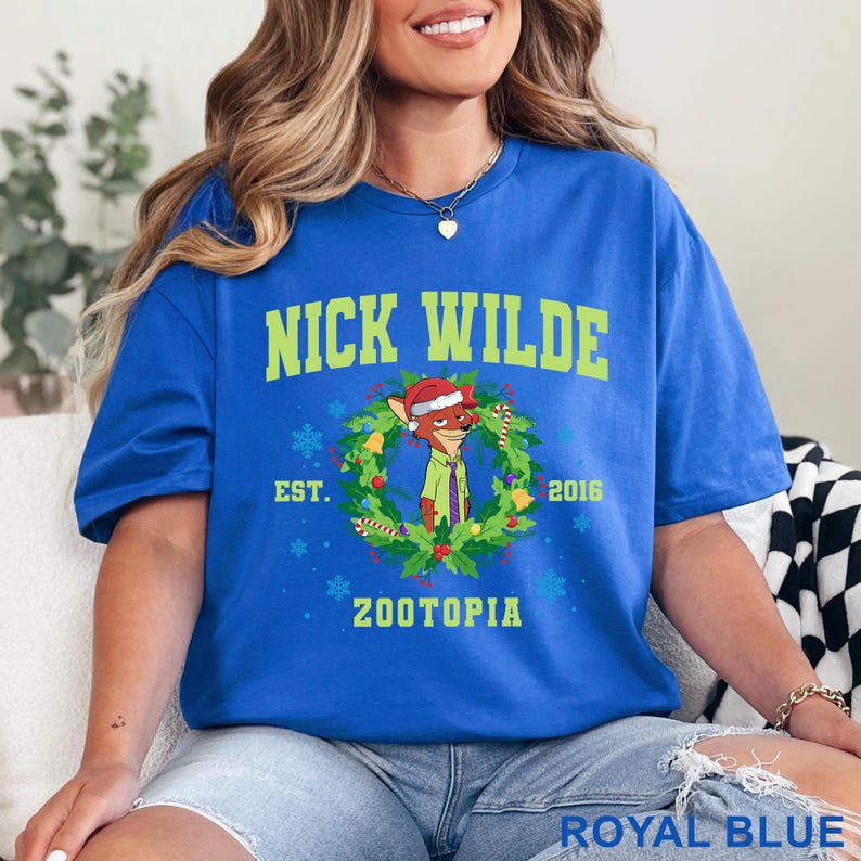 以下が含まれることがあります： ロイヤルブルーのTシャツ。緑色の「NICK WILDE」の文字の上に、クリスマスリースの中のニック・ワイルドの漫画の画像があります。シャツには「EST. 2016 ZOOTOPIA」の文字も含まれています。