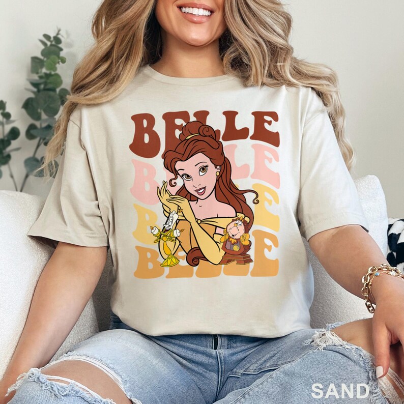 May include: Sand-colored t-shirt featuring a graphic of Belle from Beauty and the Beast. The design includes the word "BELLE" in layered retro fonts, with Belle holding Lumiere and Chip. The shirt is a relaxed fit.