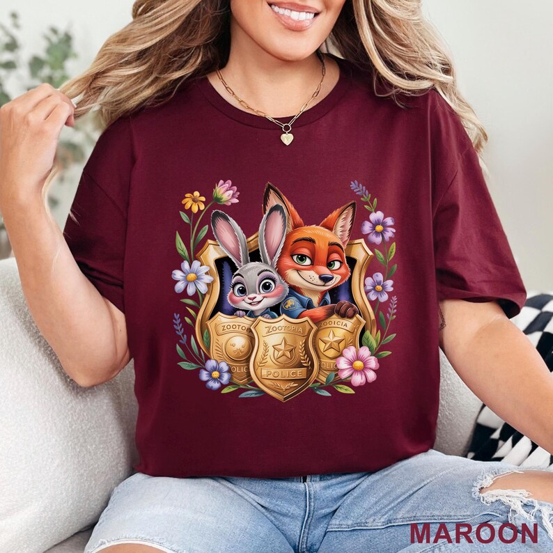 Puede incluir: Camiseta granate con una imagen de dibujos animados de Judy Hopps y Nick Wilde de Zootopia dentro de una insignia dorada, rodeada de flores. La palabra "MAROON" est&aacute; impresa en la parte inferior.