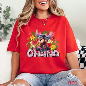 May include: Red t-shirt featuring a graphic of a blue alien character with a floral lei and surrounded by colorful flowers. The word "OHANA" is displayed in a floral font. The shirt is a solid red color.
