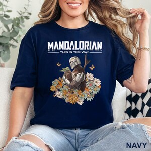 May include: Navy blue t-shirt featuring the text "MANDALORIAN - THIS IS THE WAY" in white. The graphic depicts the Mandalorian and Grogu surrounded by flowers and butterflies. The shirt is a short-sleeved crew neck.