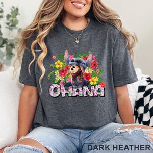 May include: Dark heather gray t-shirt featuring a colorful graphic of a blue cartoon character with a floral lei and surrounded by tropical flowers. The word "OHANA" is displayed in a floral font. The text "DARK HEATHER" is at the bottom.