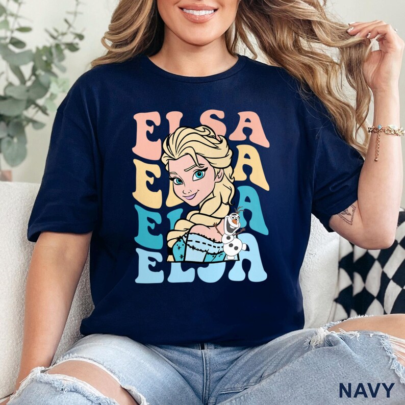 May include: Navy blue t-shirt featuring a colorful Elsa graphic. The design includes the name 'ELSA' stacked vertically in pastel colors, with an illustration of Elsa from Frozen. The character is blonde with a braid, and a small Olaf figure is also present.