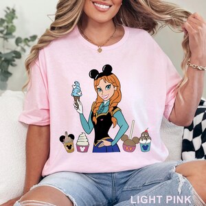 May include: Light pink t-shirt featuring a cartoon illustration of a character holding a blue ice cream cone, with other ice cream treats. The character has red hair and is wearing a black dress. The text "LIGHT PINK" is at the bottom.