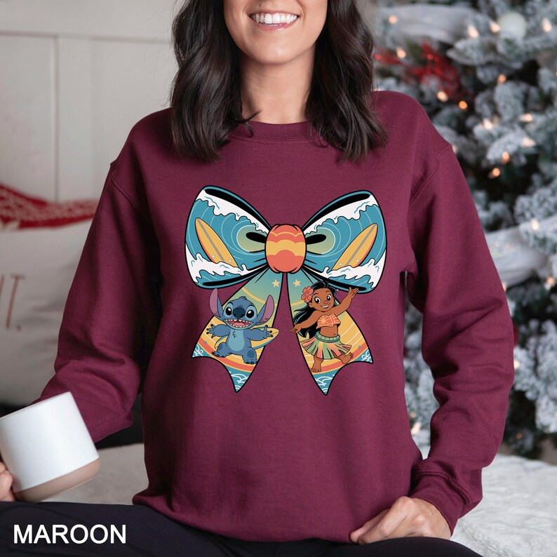 Puede incluir: Sudadera granate con un gran dise&ntilde;o de lazo con personajes de dibujos animados y tablas de surf. El lazo tiene una combinaci&oacute;n de colores turquesa y amarillo con un patr&oacute;n de olas. La palabra "MAROON" est&aacute; impresa en la parte inferior de la imagen.