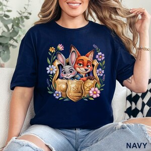 Puede incluir: Camiseta azul marino con un gr&aacute;fico colorido de Judy Hopps y Nick Wilde de Zootopia, enmarcado por flores y una placa de polic&iacute;a. La camiseta tiene mangas cortas y cuello redondo.