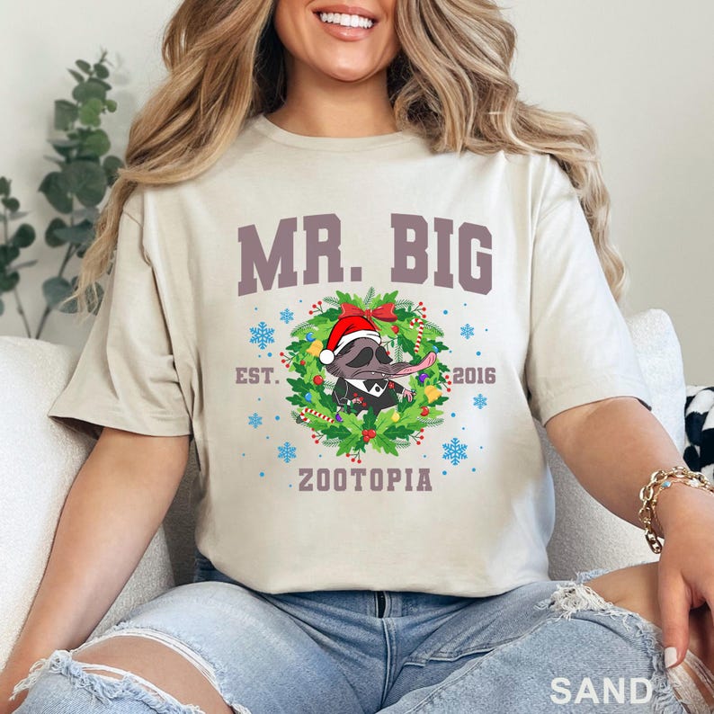 以下が含まれることがあります： 砂色のTシャツ。テキスト「MR. BIG」の下に、サンタ帽とタキシードを着たアライグマのグラフィックがあり、クリスマスのリースで囲まれています。「EST. 2016」と「ZOOTOPIA」の文字も表示されています。