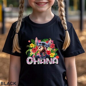 May include: Black t-shirt featuring a colorful graphic of a blue alien character surrounded by tropical flowers. The word "OHANA" is displayed in a floral font. The shirt is a short-sleeved design.
