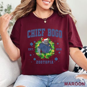 以下が含まれることがあります： 青色の「CHIEF BOGO」の文字がプリントされたマルーン色のTシャツ。サンタ帽をかぶった警察官の制服を着た漫画の雄牛が、リースに囲まれています。Tシャツには「ZOOTOPIA」と「2016」の文字も含まれています。