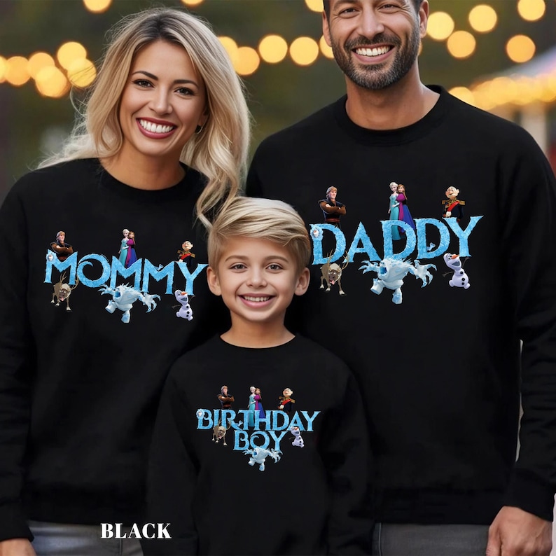 Op de afbeelding: Zwarte sweatshirts met de woorden "MOMMY", "DADDY" en "BIRTHDAY BOY" in blauw, samen met cartoonfiguren. Een gezin van drie draagt de sweatshirts. Het woord "BLACK" staat onderaan de afbeelding.
