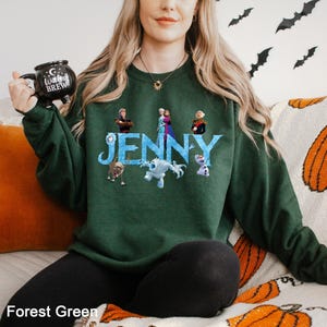 Op de afbeelding: Boszwarte groene sweatshirt met de naam "Jenny" in blauw, met personages uit een populaire animatiefilm. De sweatshirt is effen van kleur en heeft een ronde hals. Een zwarte mok met de woorden "Witches Brew" wordt in de linkerhand gehouden.