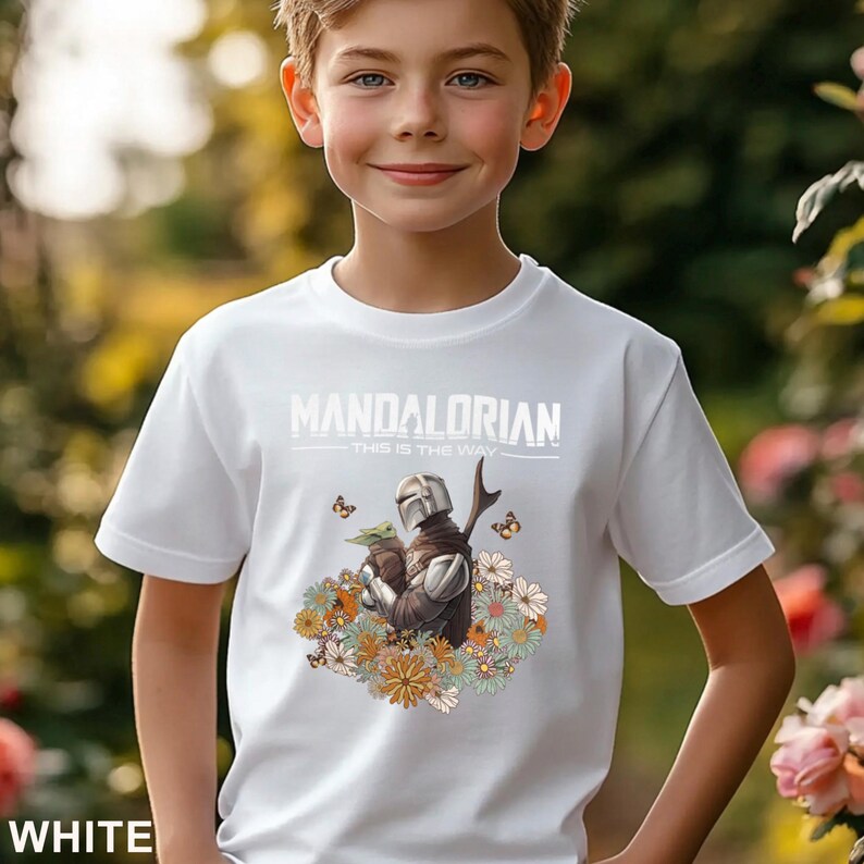 May include: White t-shirt featuring a graphic of The Mandalorian and Grogu surrounded by flowers and butterflies. The text "MANDALORIAN THIS IS THE WAY" is printed across the top. The shirt is made of a soft material.