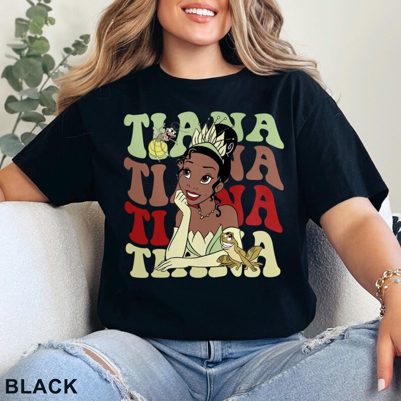 May include: Black t-shirt featuring a graphic of Princess Tiana with the word "Tiana" in green, brown, and red retro lettering. The design includes a cartoon frog and a small cartoon character. The word "BLACK" is at the bottom.