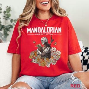 May include: Red t-shirt featuring the text "MANDALORIAN" above the phrase "THIS IS THE WAY." The graphic depicts the Mandalorian and Grogu surrounded by flowers and butterflies. The shirt is a vibrant red color.