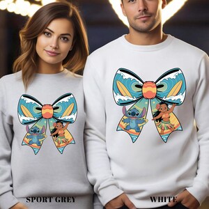 Puede incluir: Sudaderas deportivas grises y blancas con un gran dise&ntilde;o de lazo con los personajes de dibujos animados Stitch y Lilo. El lazo tiene olas azules, tablas de surf amarillas y un centro naranja. El texto "SPORT GREY" y "WHITE" est&aacute; en la parte inferior.