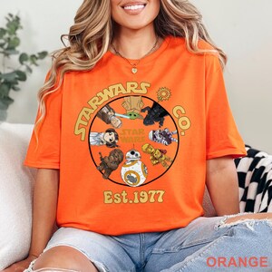 Peut inclure: T-shirt orange vif avec un motif Star Wars. Le graphisme circulaire pr&eacute;sente divers personnages et le texte "STARWARS CO. Est. 1977". Les personnages incluent Yoda, Dark Vador et BB-8. Le t-shirt est de couleur orange vif.