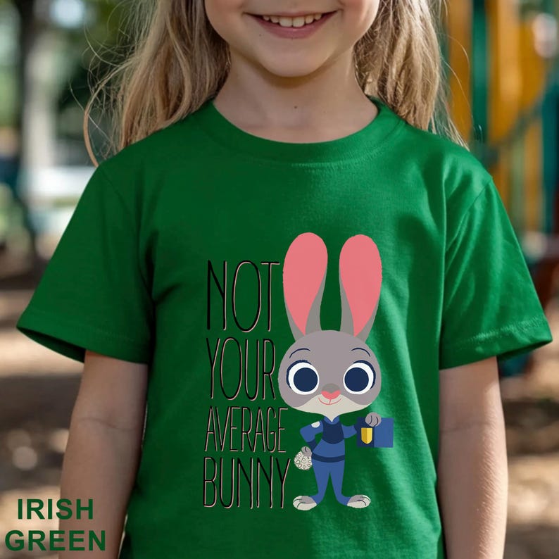 Puede incluir: Camiseta verde irland&eacute;s para ni&ntilde;os con un personaje de conejo de dibujos animados con uniforme azul, sosteniendo un malet&iacute;n y una bolsa de zanahorias. El texto "NOT YOUR AVERAGE BUNNY" est&aacute; impreso en la camiseta.