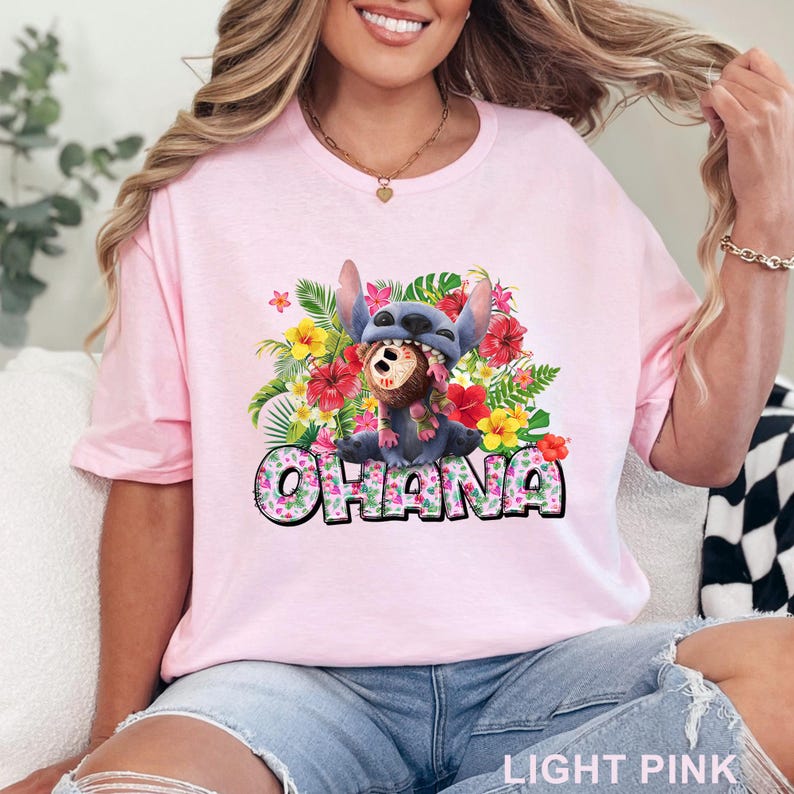 May include: Light pink t-shirt with a colorful graphic of Stitch wearing a Hawaiian lei and a hat. The word 'OHANA' is printed in a floral font below the character. The shirt is made of soft fabric and has a crew neck. The overall theme is Hawaiian and Disney.